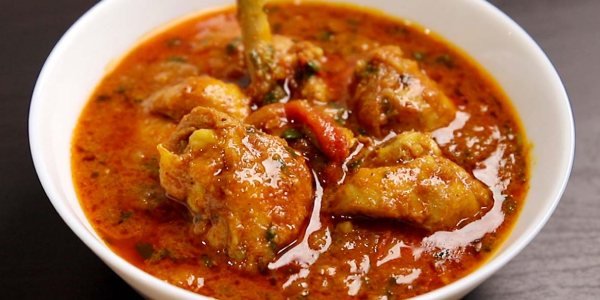 A close-up, appetizing shot of a rich Indian chicken curry served in a white bowl. The vibrant orange-red gravy is filled with tender-looking chicken pieces, including a drumstick, and is flecked with herbs. The bowl is set on a dark surface.