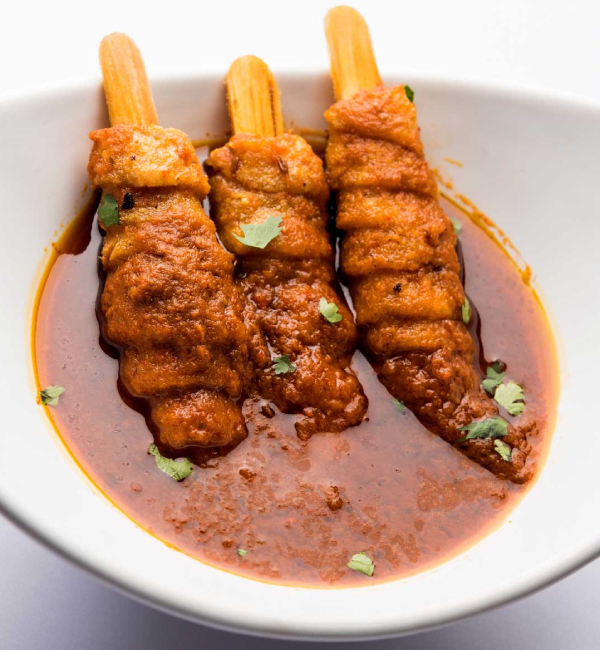 A close-up shot of an Indian dish, likely Soya Chaap Curry, in a white bowl. Three pieces of savory soya chaap, served on popsicle-like sticks, are arranged in a rich, reddish-brown gravy and garnished with fresh cilantro.