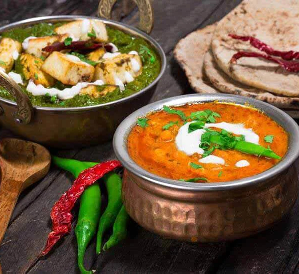 An appetizing close-up of a North Indian vegetarian meal on a rustic wooden table. The spread includes a bowl of Palak Paneer (spinach and cheese curry) and a hammered copper bowl of what appears to be Dal Tadka (lentil curry), both garnished with cream. The meal is served with whole wheat rotis and decorated with fresh green and red chili peppers.