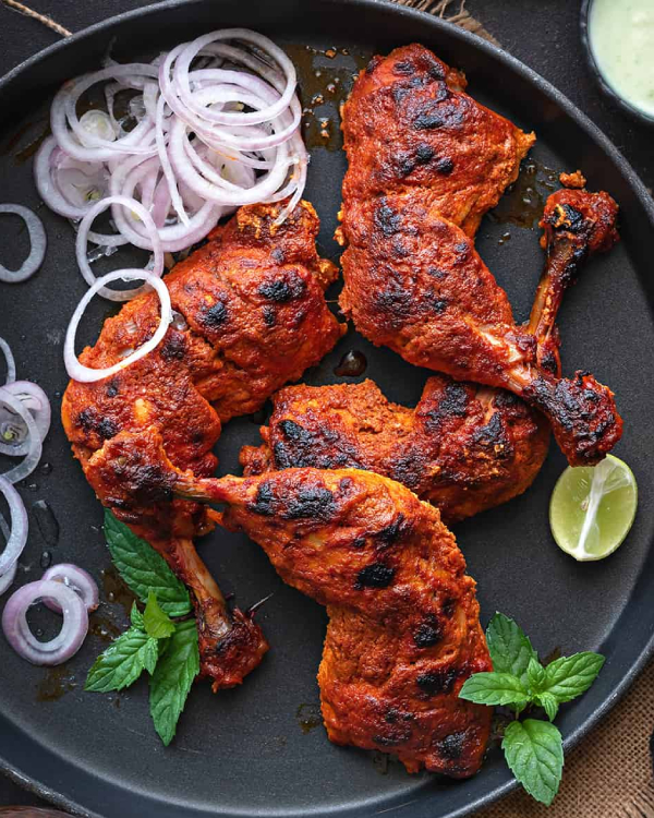 A top-down shot of vibrant red tandoori chicken served on a round black plate. Four charred chicken leg and thigh quarters are arranged with sliced red onions, fresh mint sprigs, and a lime wedge. A small bowl of green chutney is visible in the top right corner.