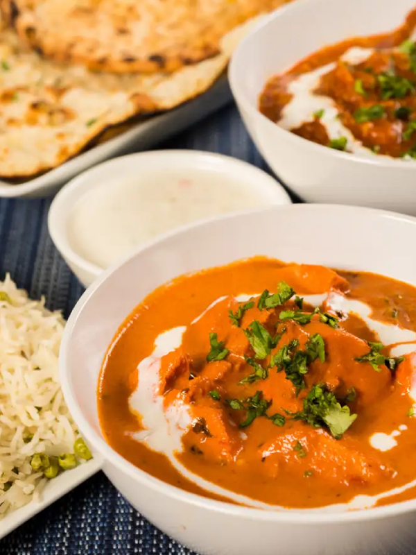 A close-up, appetizing shot of an Indian meal, with a white bowl of butter chicken in the foreground. The creamy, orange curry is garnished with fresh cilantro and a swirl of cream. In the soft-focus background, there is another bowl of curry, naan bread, raita, and basmati rice with peas, all arranged on a blue placemat.