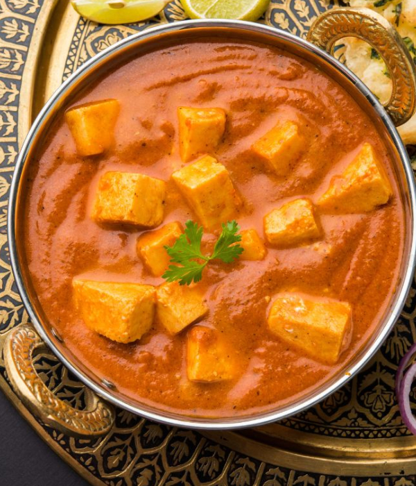 A close-up, top-down shot of paneer butter masala, a popular Indian vegetarian dish. The creamy, orange-colored curry with cubes of paneer cheese is served in a traditional metal karahi with decorative golden handles. The karahi is placed on an ornate gold and black tray, with lime wedges and naan bread visible in the background.