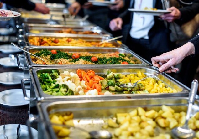 A buffet line with several chafing dishes filled with different foods. People are serving themselves from the dishes. In the foreground, there are trays of cooked vegetables like broccoli, cauliflower, carrots, and Brussels sprouts, as well as roasted potatoes. Further down the line, there appear to be rice dishes and possibly pasta. Several hands are visible holding serving utensils or plates. The setting suggests a catered event or a large gathering.
