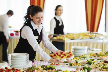 A smiling female caterer in a black vest and white shirt is placing a platter of small fruit tarts onto a table laden with an extensive buffet spread. Behind her, another caterer pushes a cart with more food, and a man in a white shirt is partially visible in the background. The room has warm-toned curtains and a celebratory atmosphere, suggesting an event.