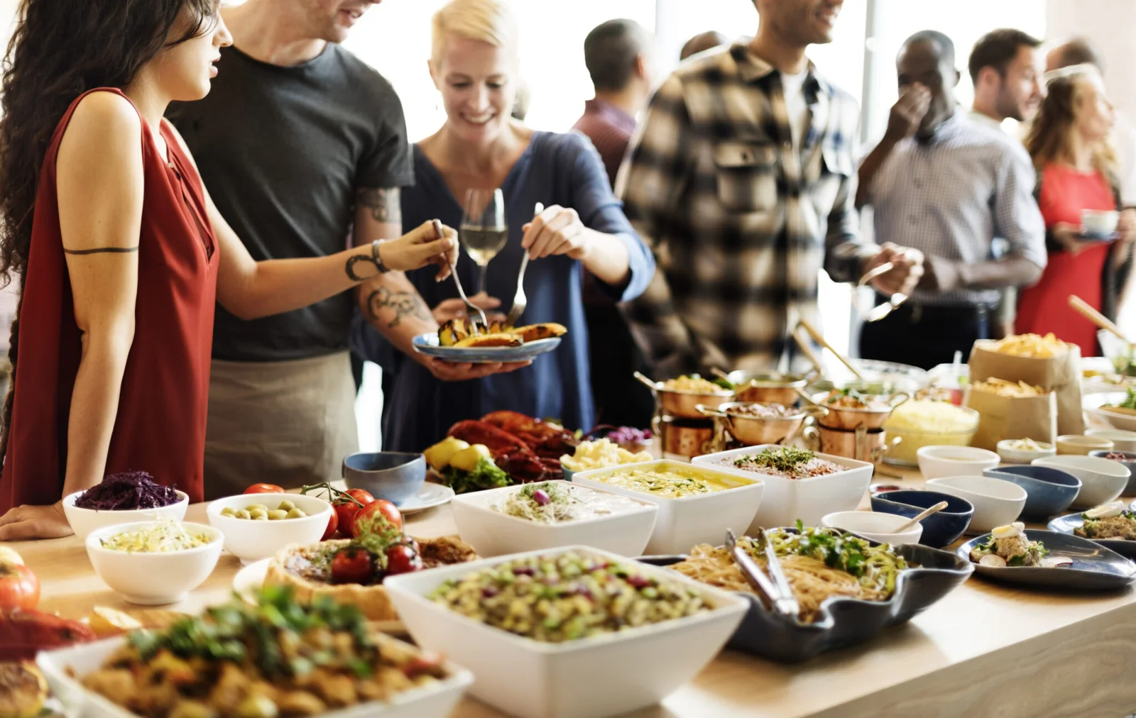 A group of diverse people are gathered around a vibrant buffet table, serving themselves food. In the foreground, numerous bowls and platters are filled with various colorful dishes, including salads, pasta, and what appears to be curries or stews in small copper pots. A woman in a red top and a man in a black t-shirt are selecting food, while a smiling blonde woman in a blue top is also serving herself. Other guests are visible in the background, enjoying the event. The atmosphere appears lively and social.