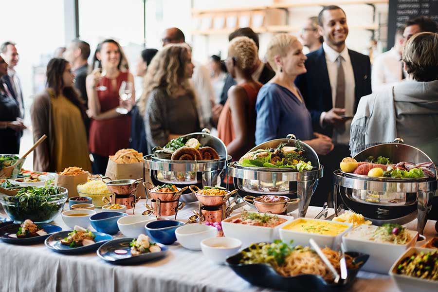 A vibrant scene showing a large catering buffet spread with various dishes in chafing dishes and bowls, with elegantly dressed people mingling in the background.
